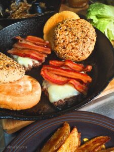 a cast iron pan with two venison burgers, topped with melted cheese and bacon slices and toasted buns. A pan of caramelized onions is in the background, with romaine lettuce and a plate of oven fries.