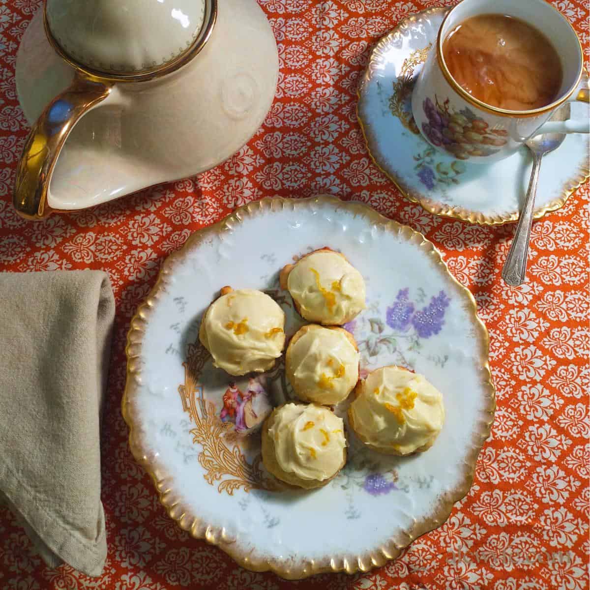 a fancy gilded antique china place setting with a teapot, a cup of tea with cream, a folded cloth napkin, and a plate of frosted creamsicle cookies, garnished with orange zest.