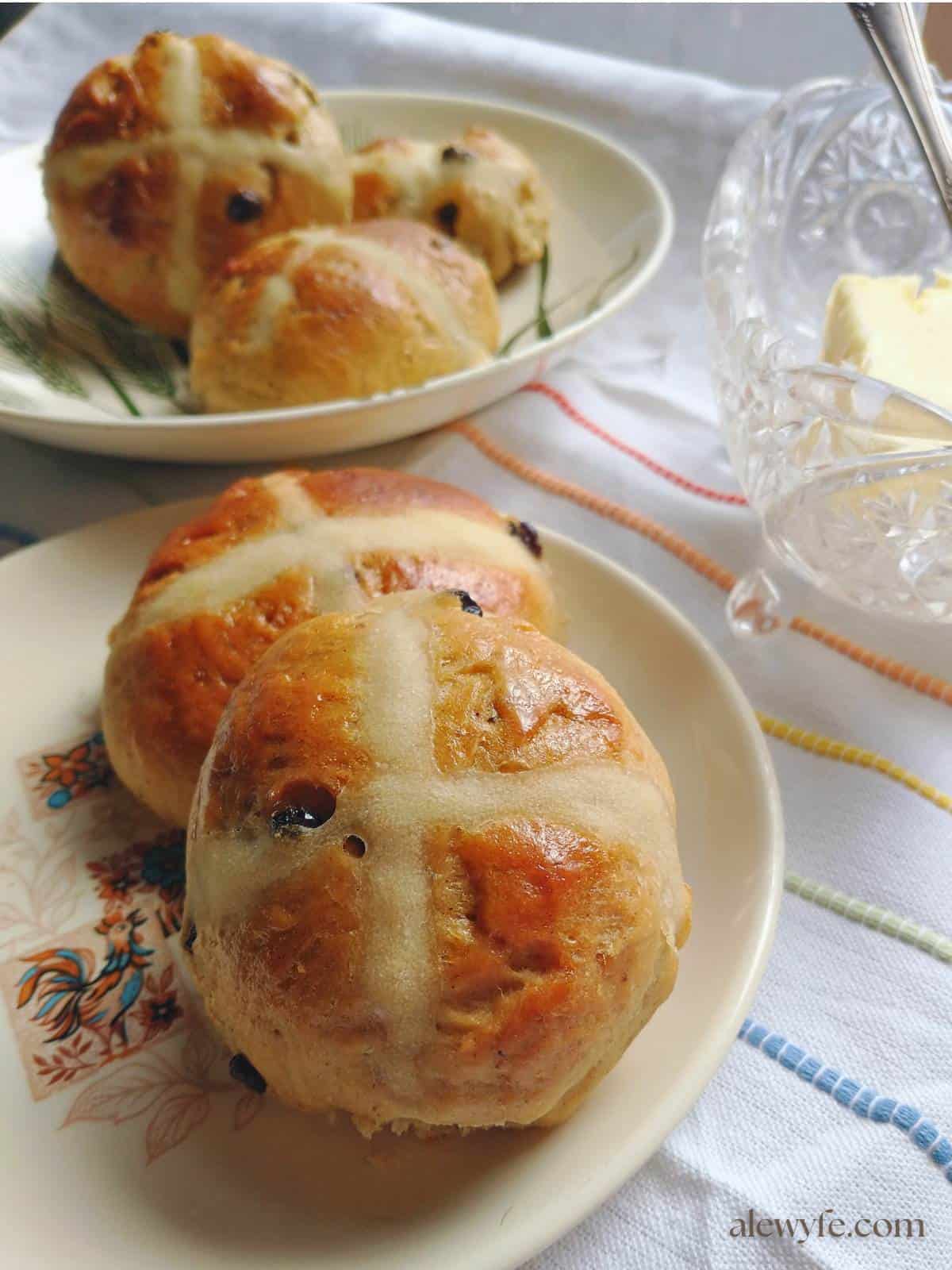 plates with hot cross buns on a white tea towel with colorful striped embroidery next to a crystal butter dish.