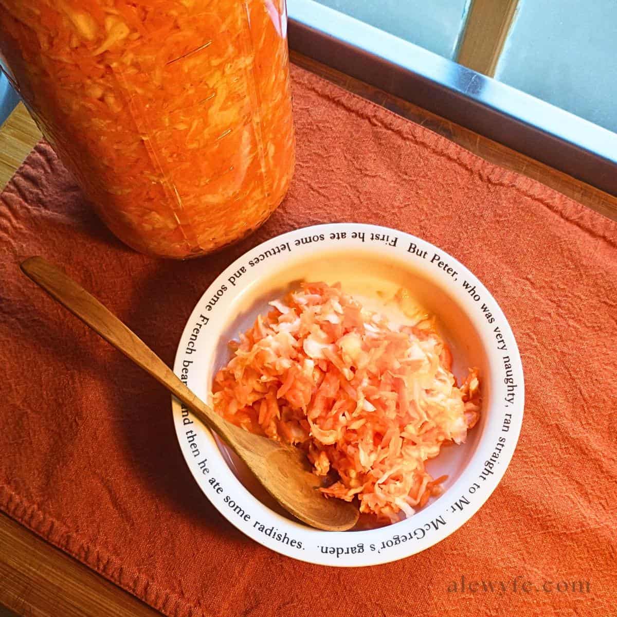 top-down photo of a bowl of ginger carrot-cabbage slaw in a bowl with Beatrix Potter quotes from Peter Rabbit on the rim, and a small wooden spoon. There's a large mason jar filled with the homemade kraut.