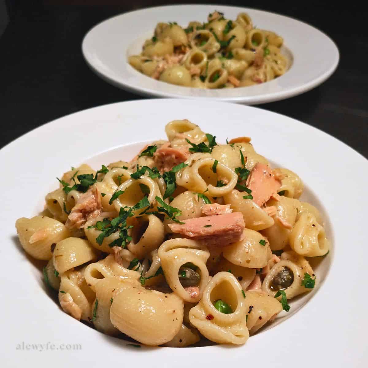 two white wide-rimmed bowls of spicy tuna pasta made with lumache, peas, and capers with bright green parsley.