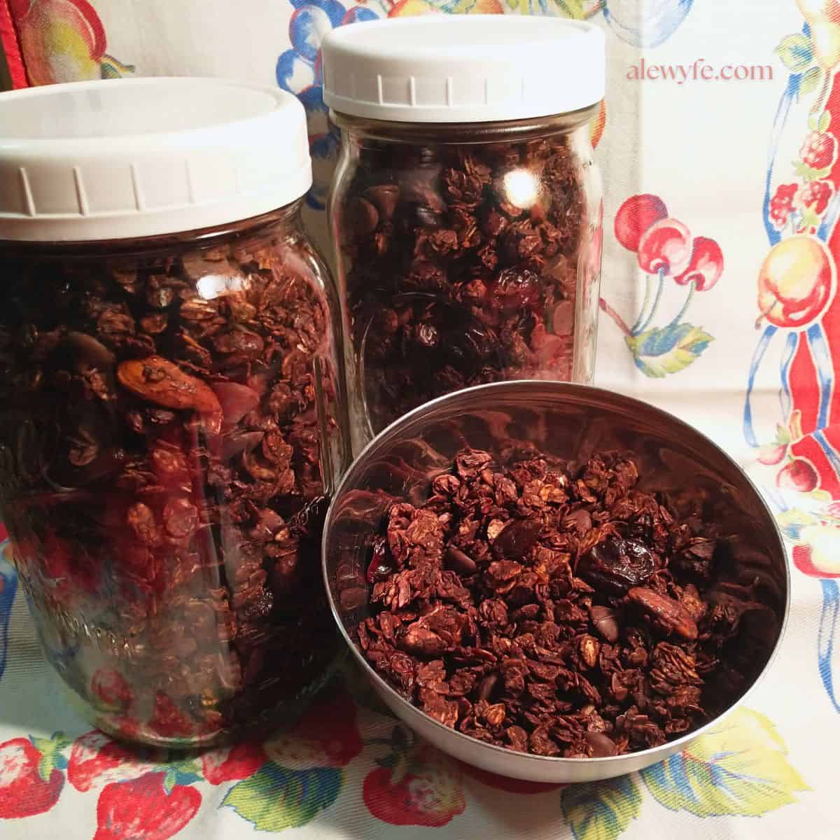 two quart jars and a small bowl of chocolate cherry almond granola, on a vintage fruit printed tea towel.