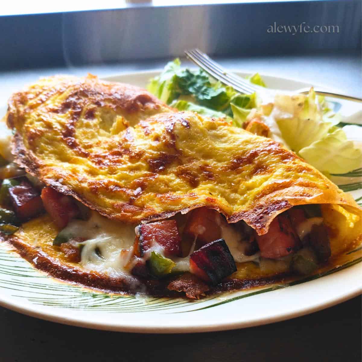side angle view of a plated denver omelette with golden brown edges, diced ham, peppers, and melted cheese, on a plate with a small green salad.