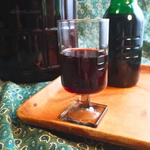 a small midcentury modern wine glass of homemade elderberry wine on a wooden tray with a green glass bottle of wine. The tray and a one gallon fermenter are on a green paisley cloth background.
