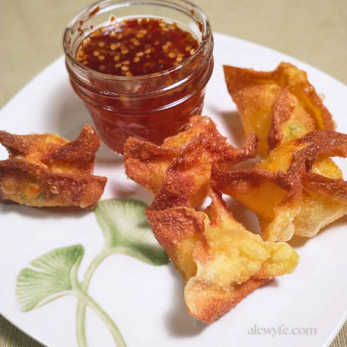 five golden fried jalapeno popper wontons on a plate decorated with a ginko leaf along with a jar of sweet chili garlic dipping sauce.