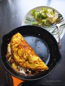 a small le creuset pan with a folded denver omelette, just before sliding the omelette onto a plate next to a bit of dressed green salad and a fork on it.