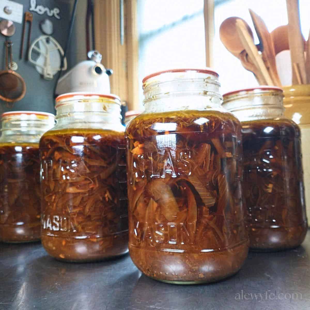canning jars of french onion soup lined up on a stainless kitchen prep table, backlit by a window.