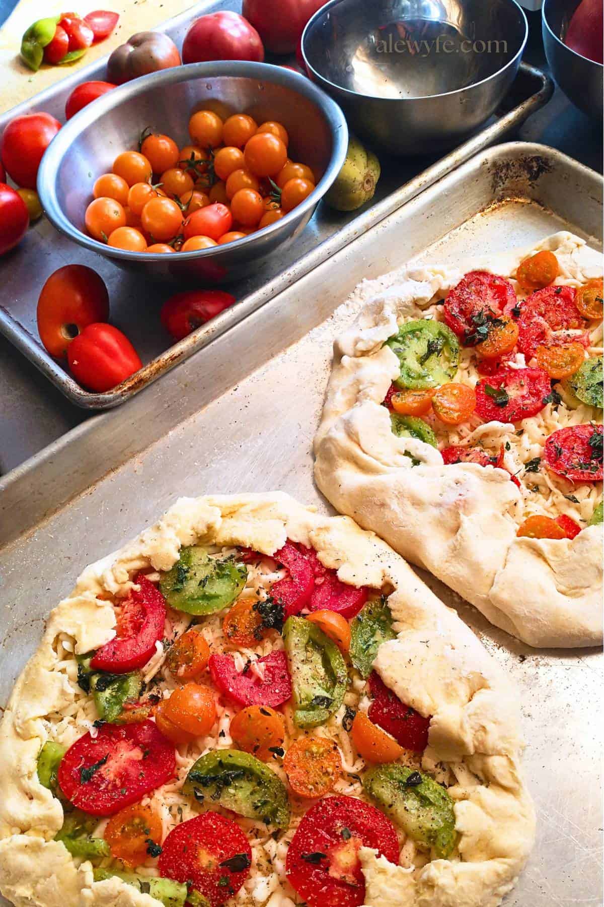 two rustic tomato tarts on a sheet pan ready to be baked, next to a tray with bowls of heirloom tomatoes. 