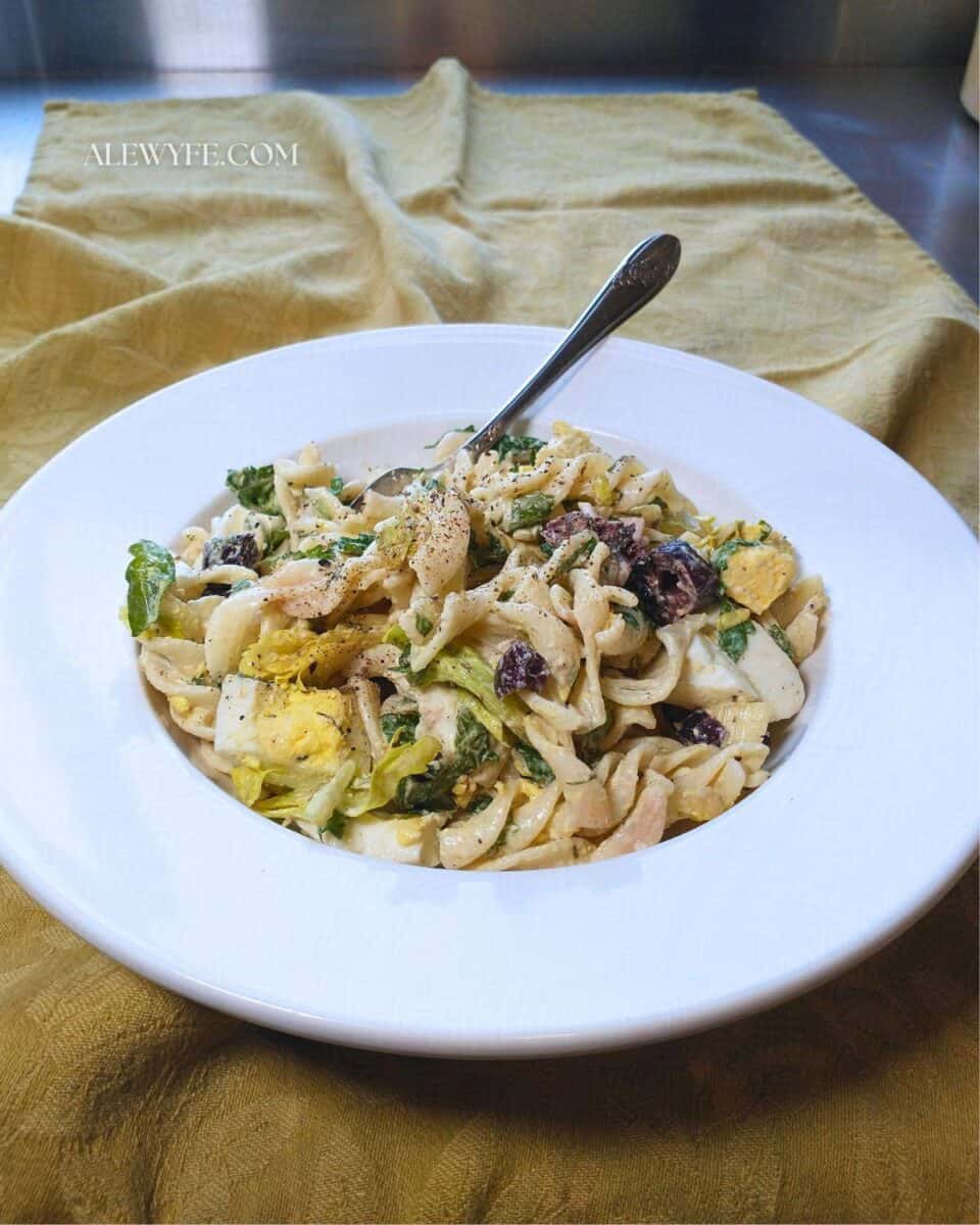 a wide rimmed white bowl of creamy pasta salad with arugula & olives.