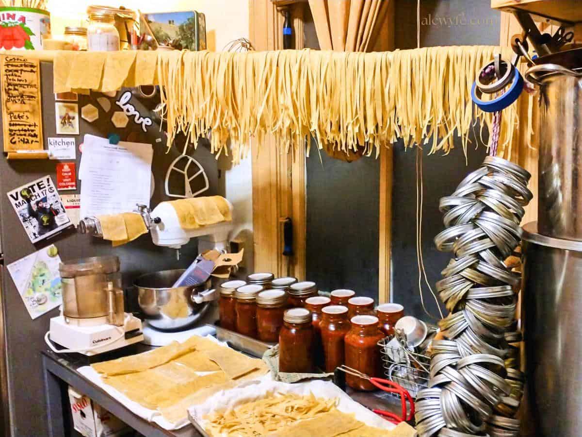 Fresh homemade pasta (egg noodle fettuccine) is drying on a bamboo pole hung between two shelves in a kitchen. Large pots and canning jar rings are in the foreground, and in the background trays of homemade pasta are drying and canned mason jars full of tomato ragu are on the counter.