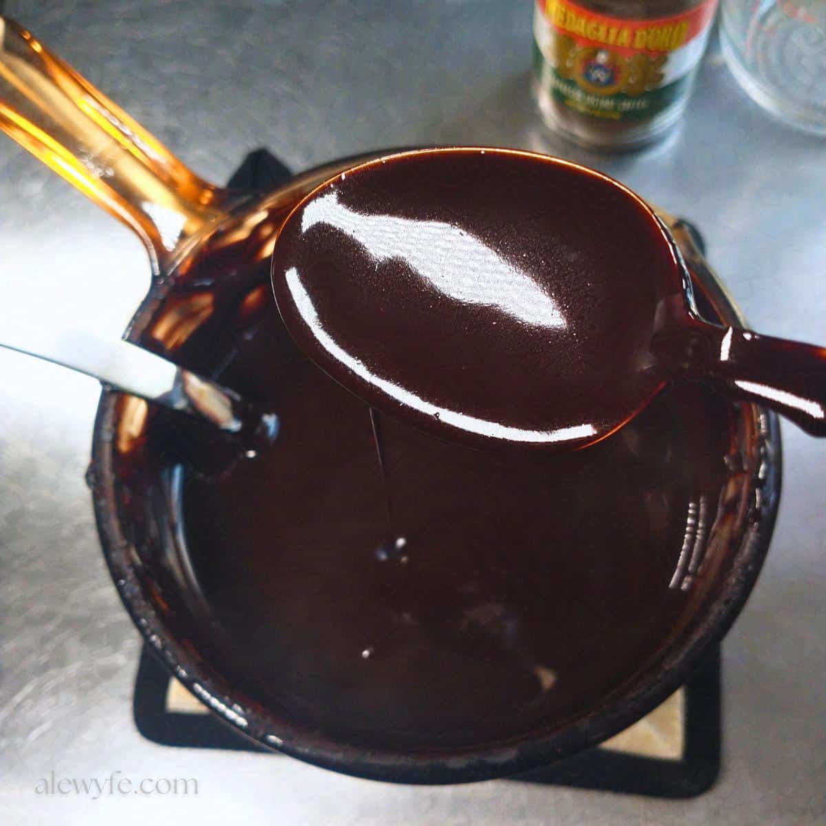 a spoon coated with homemade chocolate mocha syrup above a small saucepan filled with more of the chocolate sauce.