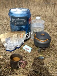 a bear canister, bottle of water, a small kettle on a trangia stove, a packet of freeze dried food, and a titanium cup of coffee on a grassy backdrop.