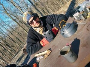 a man sits at a picnic table in the woods, holding a cup of coffee in gloved hands. 