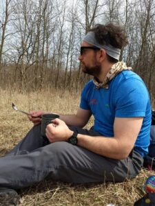 a man in sunglasses sits in a field and drinks a cup of coffee from an aluminum cup. 