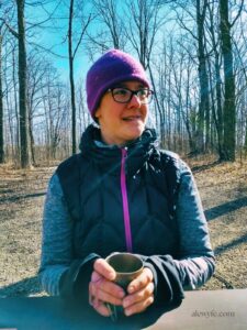 carla at a picnic table in the woods, wearing a warm hat and down vest, drinking a cup of coffee.