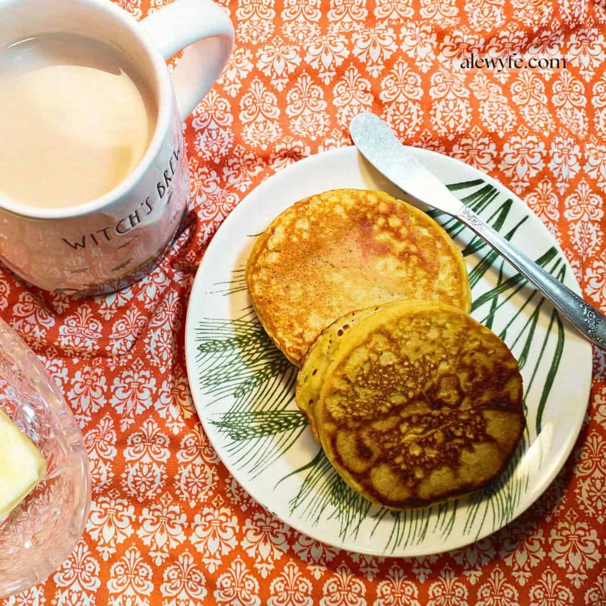 Two golden pumpkin crumpets on a small plate with a butterknife, a mug of milky tea, and a dish of butter sitting on a patterned tablecloth.