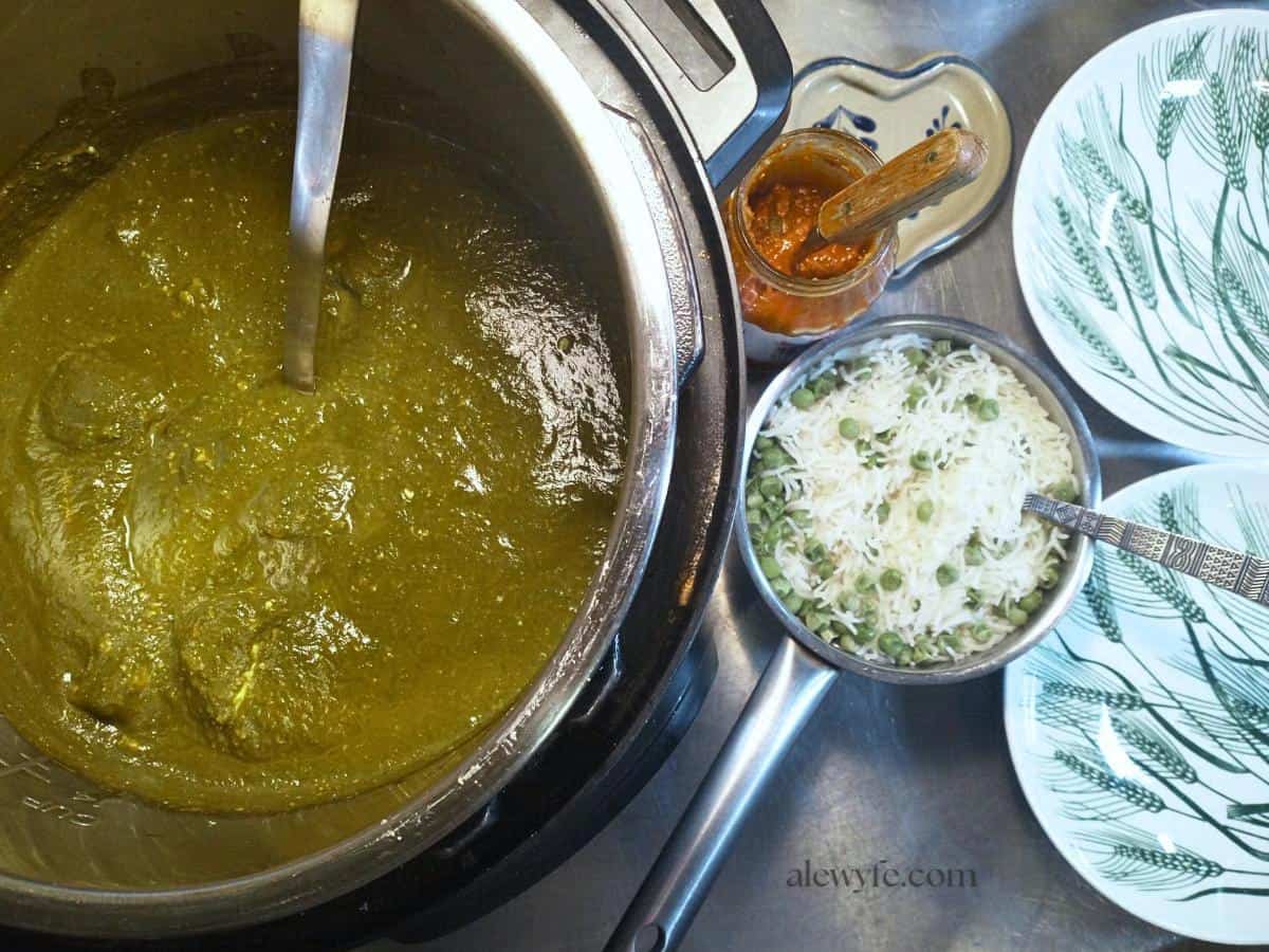 top-view of an instant pot with palak paneer curry, a pot of rice with peas, a jar of mango pickle, and two plates. 