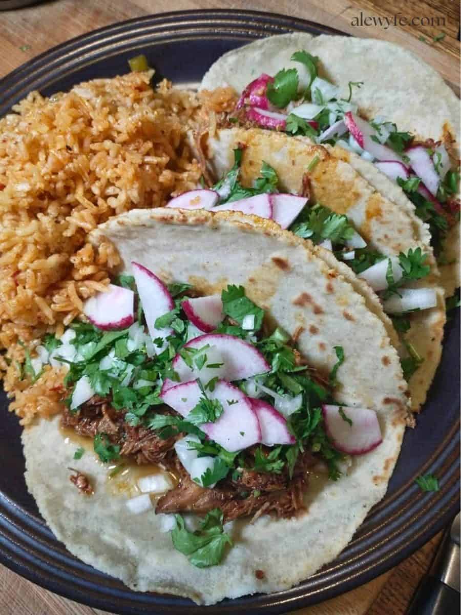 three pulled pork tacos in handmade tortillas, topped with chopped cilantro, onion, and radishes, with a side of Spanish rice. 