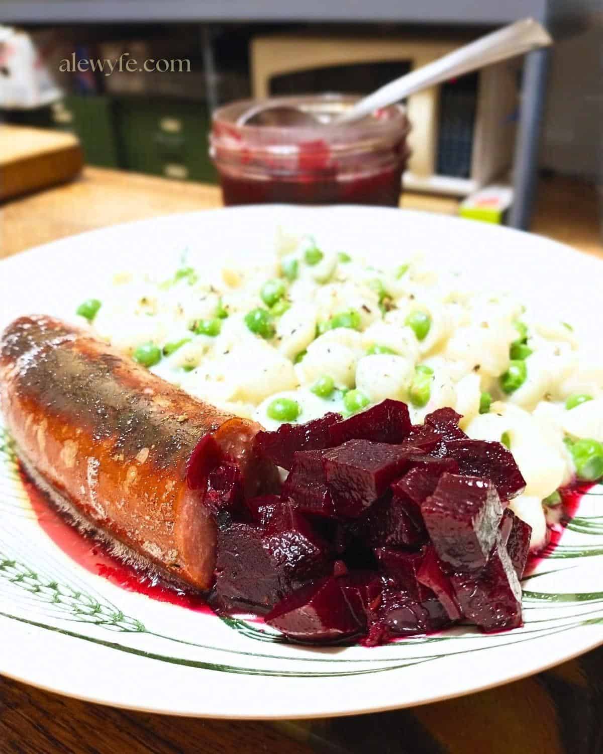 a polish kielbasa sausage link on a plate with diced beet relish and white cheddar macaroni with peas. an open jar of relish is in the background with a spoon.