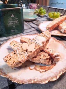 elegant plates with biscotti, drizzled with white chocolate and slivered almonds. A tin of tea in the background, Harney & Sons Victorian London Fog, with tea cups, saucers, and fruit in the background.