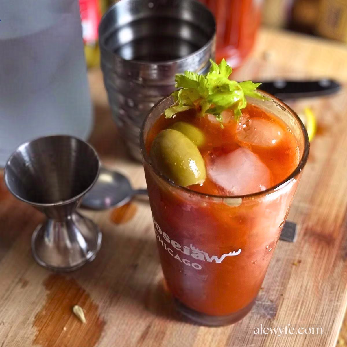 a freshly mixed bloody mary in a pint glass, with a cocktail shaker, spoon, bottle of vodka, and a shot glass in the background.