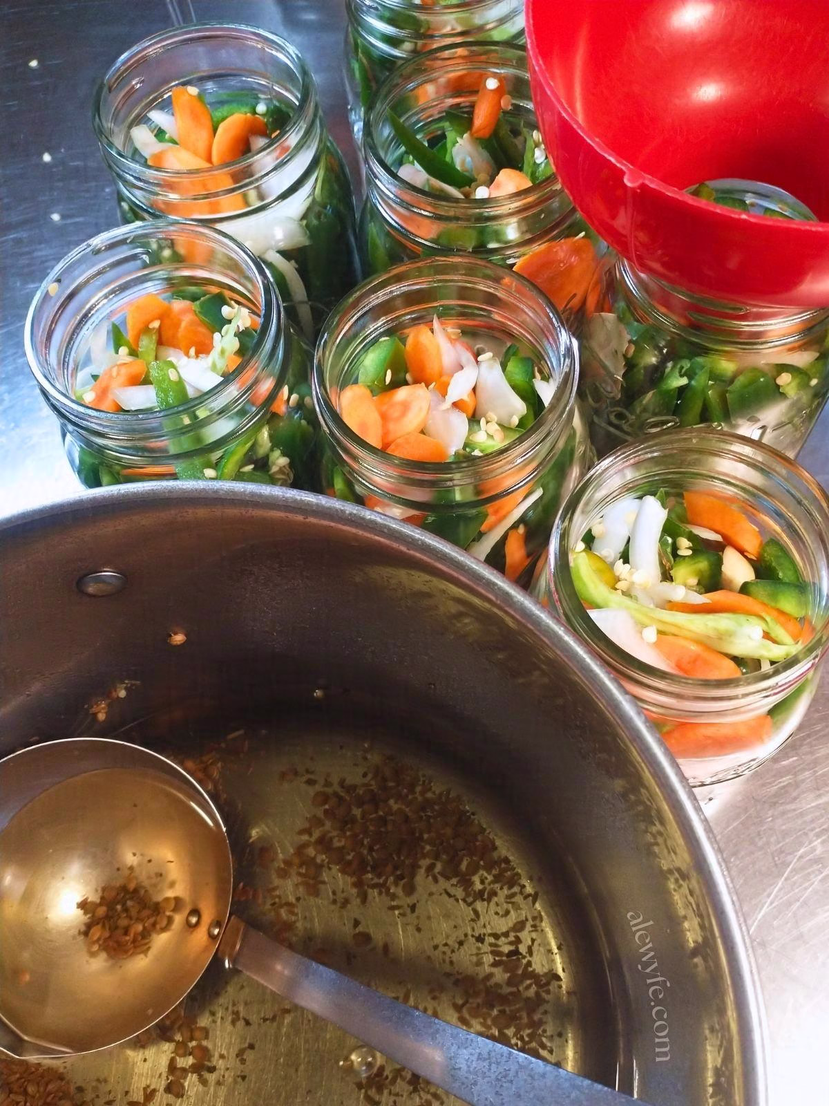 canning jars packed with strips of jalapeno peppers, onion, and sliced carrots, with a pot of pickling brine and a ladle in the foreground and a wide-mouth funnel on one of the canning jars.