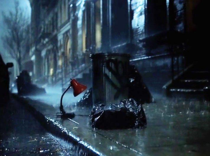 a still image from an Ikea commercial, with a red lamp outside on a city curb in the rain, next to a metal trash can and bags of trash.