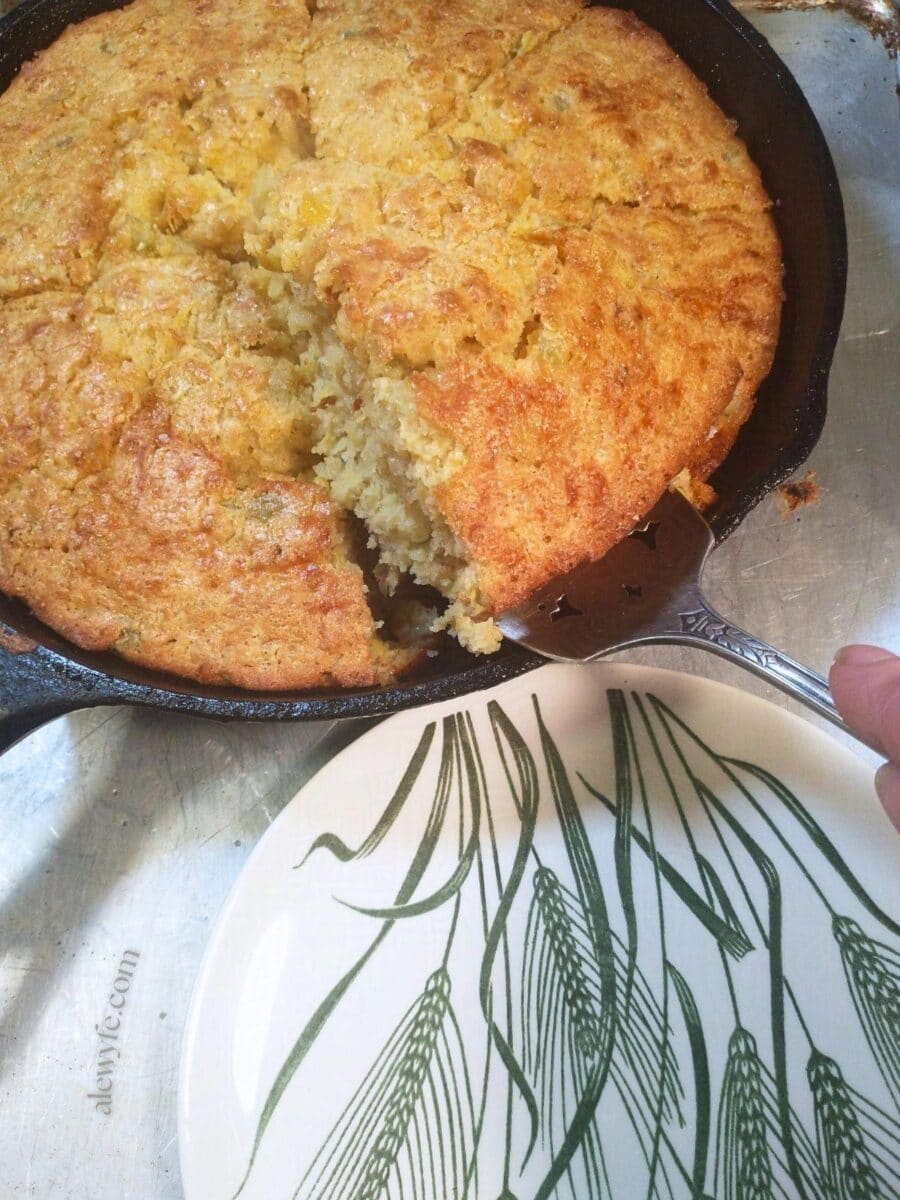 a cast iron skillet with green chile cheddar cornbread, a green and white wheat print vintage plate, and a silver pie server lifting a wedge of cornbread towards the plate.