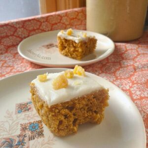 two plated slices of ginger pumpkin bars with cream cheese frosting, topped with chopped pieces of candied ginger. They're on small dessert plates resting on an orange and cream fleur de lis print tablecloth, with a vintage ceramic crock in the background. alewyfe.com watermark.