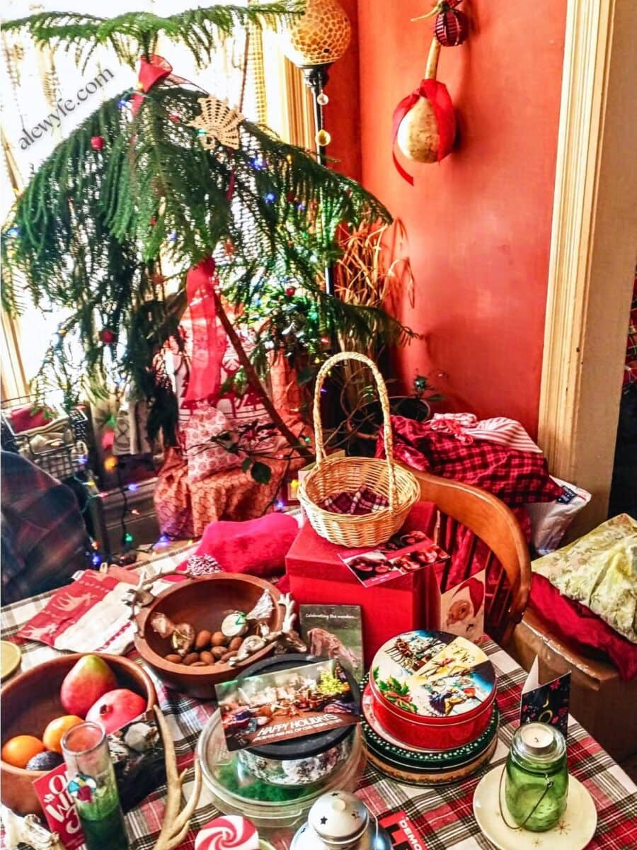 a festive table with a plaid tablecloth, vintage cookie tins, candles in colorful jars, bowls of fruit and nuts, holiday cards, and a norfolk pine tree in the background with lights and decorations. there are fabric gift bags under the tree.