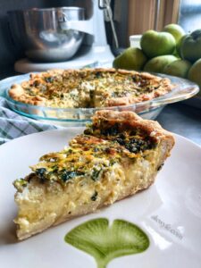 a slice of spinach, feta and artichoke quiche on a ginko-leaf printed plate with the quiche, a bowl of pears, and a mixer in the background.