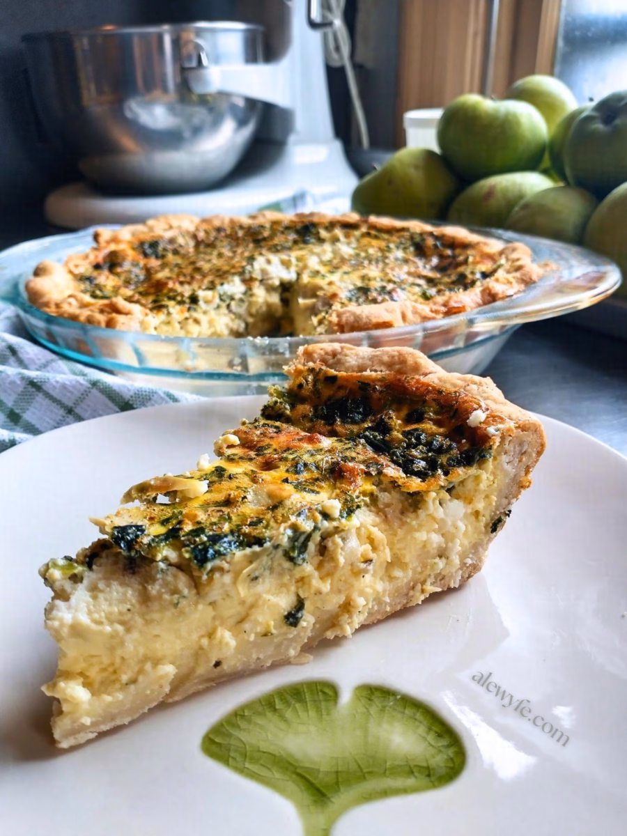a slice of spinach, feta and artichoke quiche on a ginko-leaf printed plate with the quiche, a bowl of pears, and a mixer in the background. 