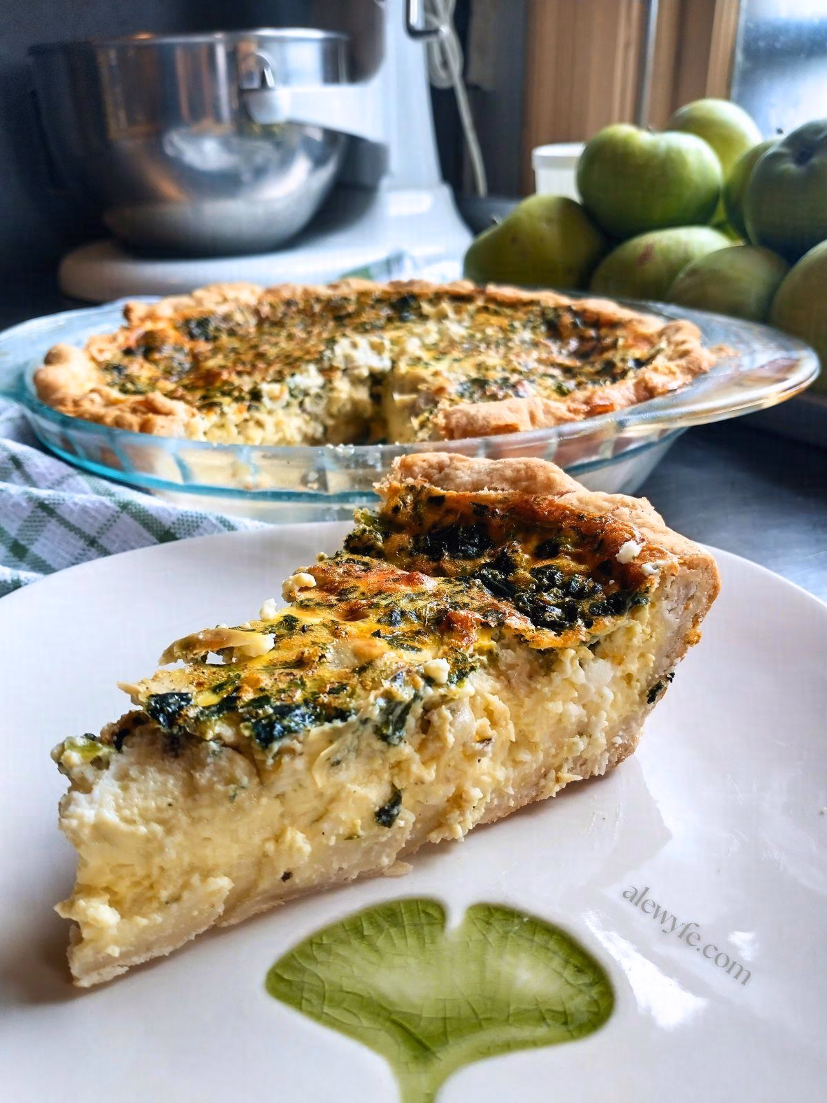 a slice of spinach, feta and artichoke quiche on a ginko-leaf printed plate with the quiche, a bowl of pears, and a mixer in the background.