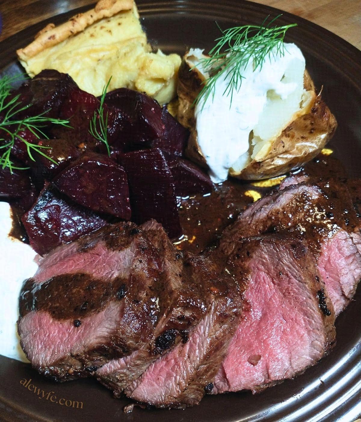 a brown ceramic plate with pan-seared rare sliced venison loin, roasted beets, yorkshire pudding, and a baked jacket potato topped with horseradish dill crema.
