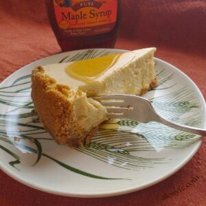a close-up of a slice of baked maple cheesecake on a plate, with a silver dessert fork, and a bottle of maple syrup in the background.