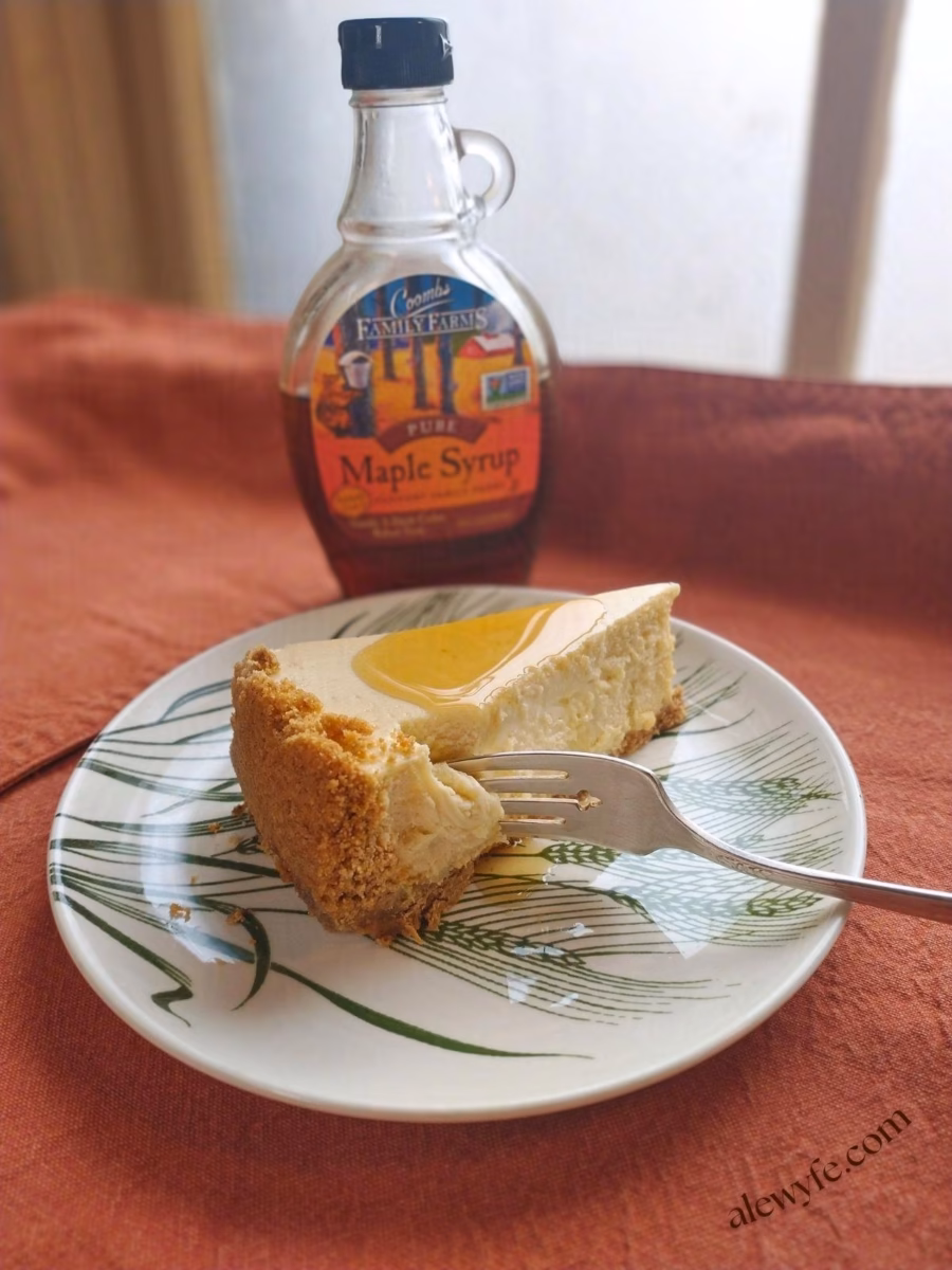 a plated slice of maple cheesecake with a drizzle of syrup on top, in front of the glass bottle of maple syrup.