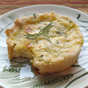 a close-up of a potato and goat cheese tartlet on a plate, garnished with fresh dill, with a bite taken out of one side.