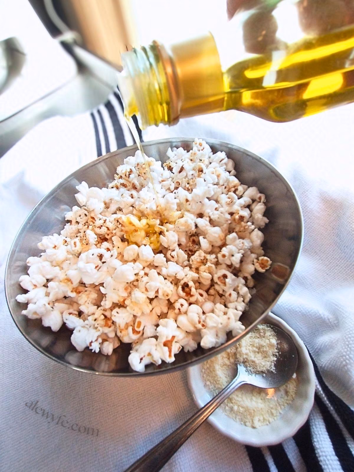 pouring truffle oil onto a bowl of seasoned homemade popcorn, with a dish of parmesan cheese and a spoon on the side.