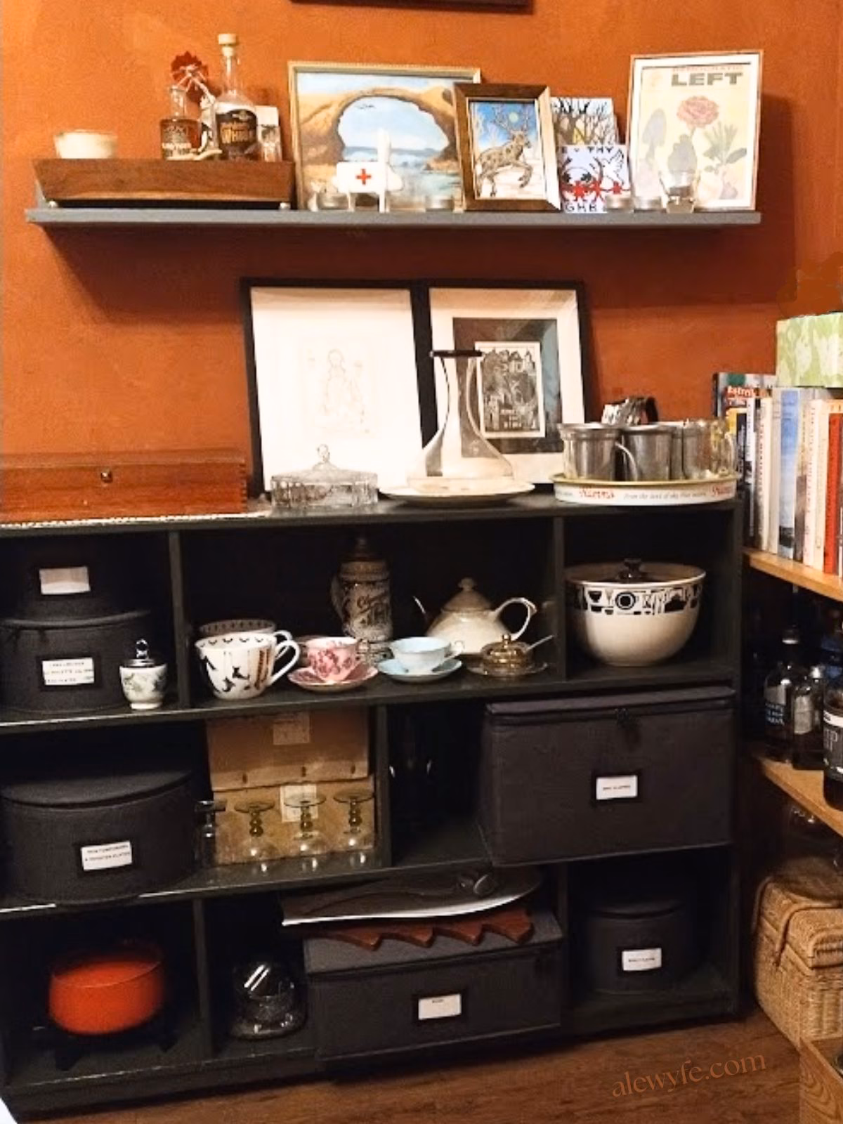 open shelving in a dining room with dark grey fabric-colored divided boxes for glassware, mugs, and dishes on dark green shelves. Other dishes are sitting on the open shelves, and books and pictures on top of the shelves.