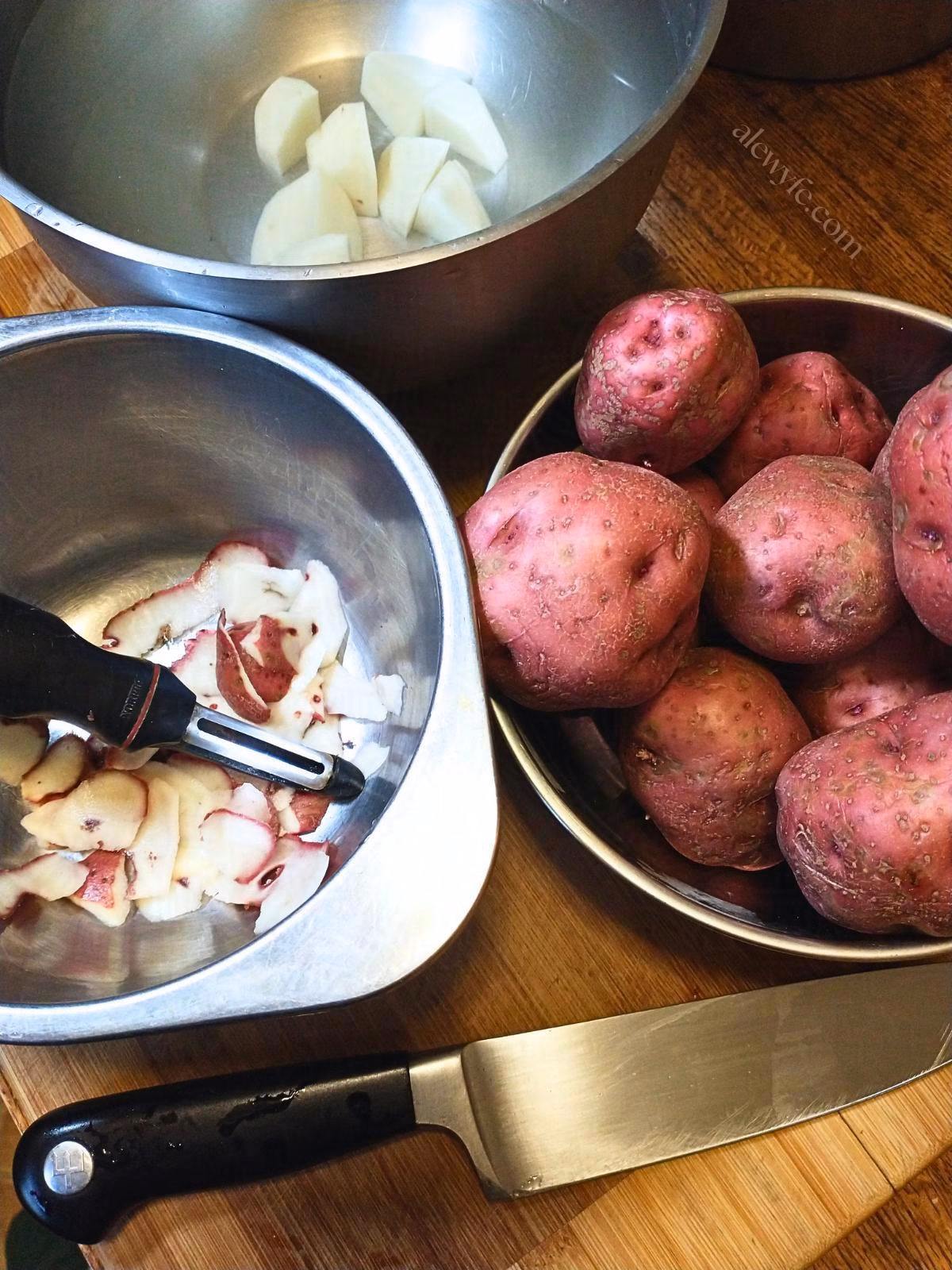 a bowl of red potatoes, with a knife and a peeler, a bowl of potato peels, and a bowl of water with diced peeled potato pieces in it.