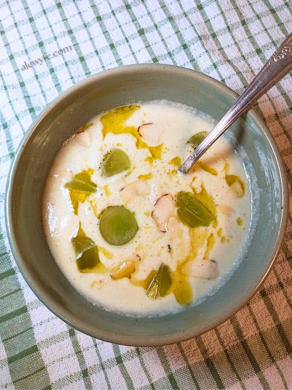a bowl of ajo blanco, a white cold cucumber soup, garnished with grapes, green olive oil, and almonds. The bowl sits on a green plaid cloth, and there is a vintage silver spoon in it.