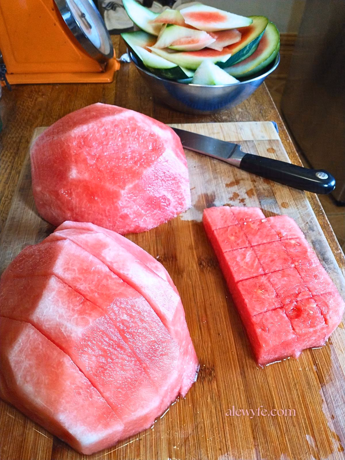 a small peeled watermelon on a bamboo cutting board. The watermelon is in various stages of slicing (one half uncut, one cut into slices and one slice cut into cubes). A bowl of melon rind is in the background along with a large knife.
