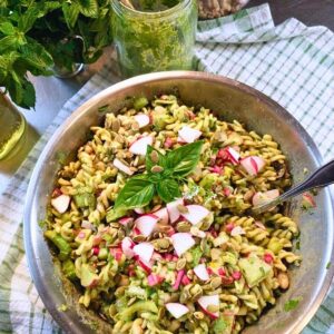 a big bowl of pasta salad dressed with chard basil walnut pesto, next to a jar of pesto. The pasta salad is garnished with fresh basil, chopped radishes, celery, white beans, and pumpkin seeds.