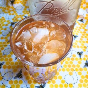 top down view into a glass of ginger peach tea on ice, in front of a mason jar full of tea, on a honeybee tea towel.