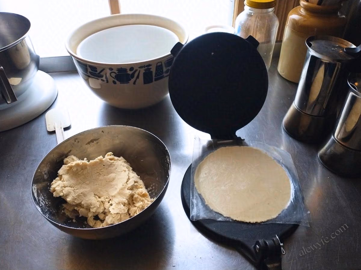 a bowl of masa next to a cast-iron tortilla press with a freshly pressed uncooked tortilla on it.