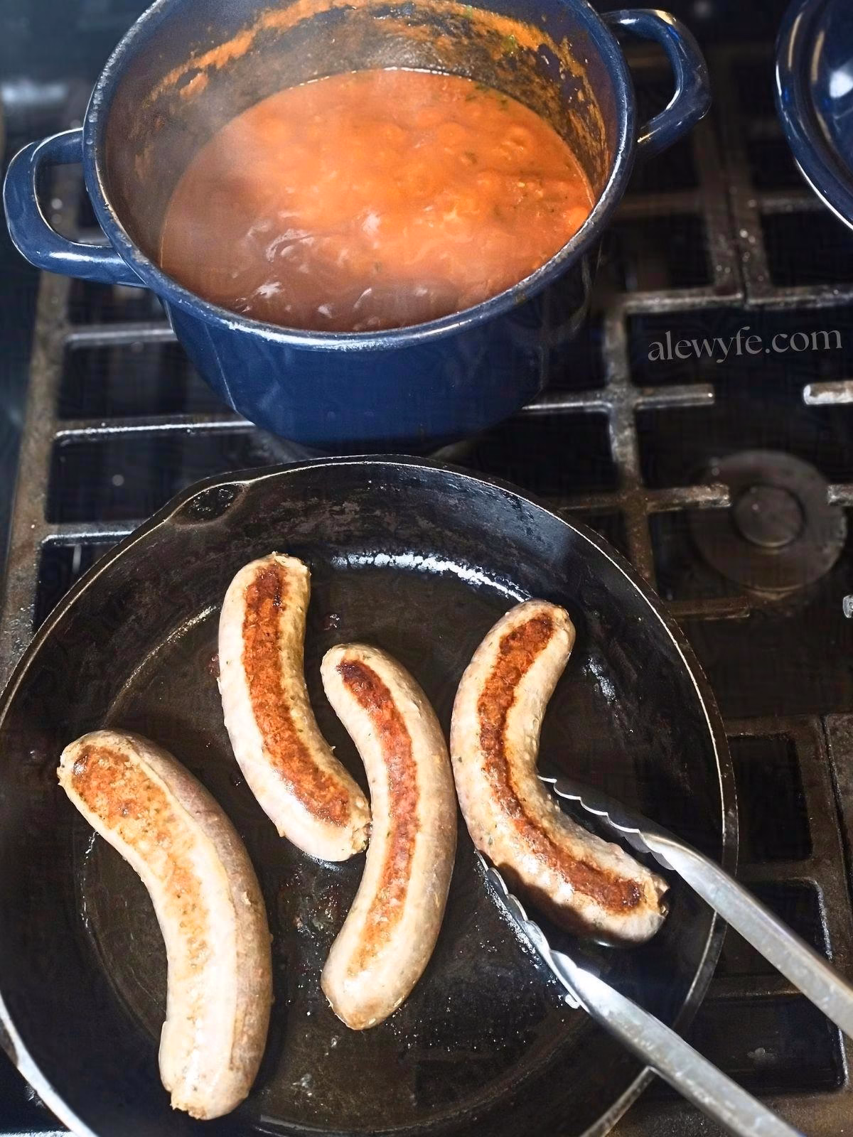 searing homemade italian sausage links in a hot cast iron pan, next to a pot of bubbling tomato sauce for braising the sausages once they are browned.